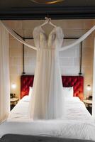 A perfect wedding dress with a lush skirt on a hanger, next to a bed with a red headboard and a white sheet, and a sconce that illuminates the bride's room. photo