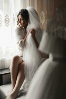 Portrait of the bride. A brunette bride in a dressing gown sits behind a dress mannequin, poses, holds her voluminous white veil and looks down. Gorgeous make-up and hair. Wedding photo. photo