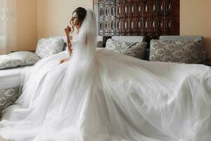 A brunette bride in a white dress poses while sitting on a gray sofa and holding her curls. Gorgeous makeup. Open shoulders. Beautiful hands. Long veil. Morning of the bride. wide angle photo