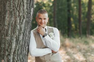 Portrait of the groom. The groom in a light gray vest is standing in the forest, leaning on a tree and touching his chin, looking into the camera lens. Wedding in nature photo