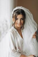 Portrait of the bride. A brunette bride in a dressing gown, posing, holding her voluminous white veil and looking into the camera lens. Gorgeous make-up and hair. Wedding photo. Beautiful bride photo