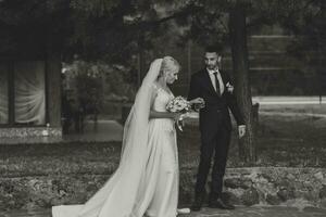 The groom in a black suit and white shirt gently touches and holds the hand of the bride in a white wedding dress, standing near a lake and stones photo