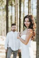 un niña con largo negro Rizado pelo soplo en el viento es en pie en un blanco seda vestido. en pie en el primavera bosque en contra el antecedentes de grande arboles foto