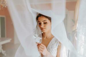 A brunette bride in a white dress holds her wedding bouquet, poses. French manicure. Open shoulders. Beautiful hands. Long veil. Morning of the bride. Details photo