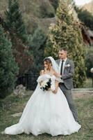 Portrait of the bride and groom in nature. The groom stands behind the bride, posing against the background of conifers. The bride in a long dress with a bouquet of roses. Stylish groom. photo