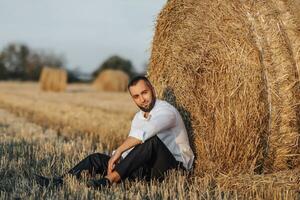 Wedding portrait photo. A stylish groom in a white shirt sits relaxed near a hay bale and looks into the lens. Bearded man. Style. photo