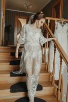 A delicate wedding dress hangs on a beautiful railing in the hallway of the house. The bride descends the steps in a delicate transparent petticoat. Nice light. Wedding photo. A high quality photo