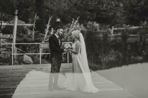 el novio en un negro traje y blanco camisa suavemente toques y sostiene el mano de el novia en un blanco Boda vestido, en pie cerca un lago y piedras foto