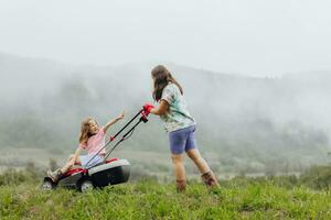 A woman in boots with her child in the form of a game mows the grass with a lawnmower in the garden against the background of mountains and fog, garden tools concept photo