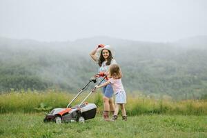 A woman in boots with her child in the form of a game mows the grass with a lawnmower in the garden against the background of mountains and fog, garden tools concept, work, nature. photo