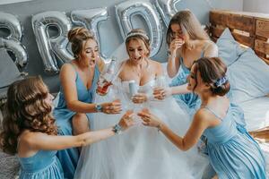 joven damas de honor en azul seda vestidos bebida champán en el de la novia habitación. hermosa mujer celebrando despedida de soltera fiesta sentado en cama y con champán. foto