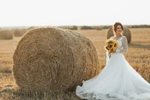 Wedding portrait. A red-haired bride in a white dress is standing near a bale of hay, holding a bouquet of sunflowers, against the background of a field. Beautiful curls. Sincere smile. Elegant dress. photo