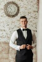 Portrait. A man in a white shirt, black bow tie and black pants and vest poses in a room. A stylish watch. Men's style. Fashion. Business photo