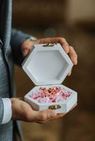 Wedding gold rings on a decorative white box with pink flowers, lying in the hands of the groom, close-up. Jewelry concept photo