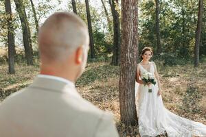 Wedding photo in nature. The bride is standing near a tree in a beautiful dress, holding a bouquet of white roses, looking at the groom. Meeting in the forest.