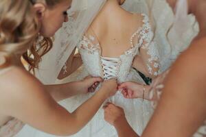 The bride's friends fasten the white voluminous dress of the bride. Rear view. Open shoulders. Beautiful hands. Long veil. Morning of the bride. photo