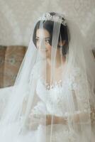 Portrait. The brunette bride is standing in a chic crown and white dress, wrapped in a veil, looking down and sniffing the groom's boutonniere. Gorgeous make-up and hair. Voluminous veil. photo
