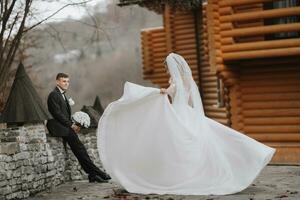 A beautiful and charming bride is circling near modern wooden houses in the park, the groom is watching. Luxurious dress with a long train. Elegant young groom photo