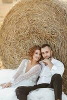 Wedding portrait. The bride and groom are sitting in each other's arms on the ground near a hay bale and looking into the lens. Red-haired bride in a long dress. Stylish groom. Summer. photo