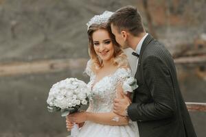 A stylish bride in a lush dress and fashionable hairstyle stands on a pier with a groom in a park near wooden houses, close-up shot photo