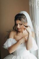 A brunette bride in a white dress holds her gorgeous curls and looks down. Open shoulders. Beautiful hands. Long veil. Morning of the bride. photo