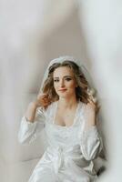 Blonde bride in a satin robe and sitting in her room next to her wedding dress, posing. Beautiful hair and make-up, open bust. Wedding portrait. photo