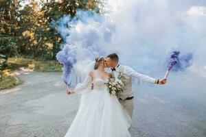 In their hands, the newlyweds hold multi-colored smoke bombs of blue color. Groom and bride kiss. Wedding fun. photo