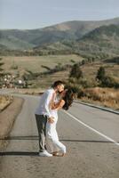 A stylish groom in a white shirt and a cute brunette bride in a white dress are hugging and kissing on an asphalt road against the background of a forest and mountains. Wedding portrait of newlyweds. photo