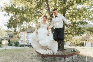 The groom in a black suit and the blonde bride in a dress and sneakers are jumping from a bench under a tree. Emotional photo of a jump. Photo session in nature. a beautiful ray of sunshine.