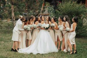 Wedding photo in nature. A brunette bride in a long white dress and her friends in nude dresses stand against a background of trees, smiling and holding their gypsophila bouquets. Young women.