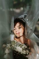 Beautiful curly brown-haired bride in a white dress poses for a photographer, standing under a veil in a beautiful dress with sleeves. Wedding photography, close-up portrait, chic hairstyle. photo