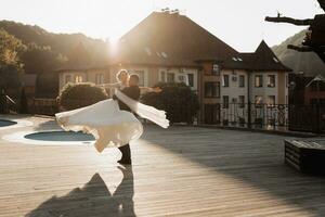 Wedding portrait. A groom in a black suit and a blonde bride dance a waltz against the background of buildings. Long dress in the air. Photo session in nature. Beautiful hair and makeup
