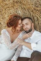 Wedding portrait. The bride and groom are sitting with their eyes closed in an embrace on the ground, near a bale of hay. Red-haired bride in a long dress. Stylish groom. Summer. photo