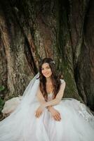 hermosa novia con largo Rizado pelo en un elegante vestido, sonriente, mirando dentro el lente debajo un grande árbol. retrato de el novia. primavera boda. natural maquillaje foto