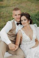 Wedding portrait of the bride and groom. Happy newlyweds are sitting gently holding hands on the green grass, posing. Stylish groom. Beautiful young bride photo