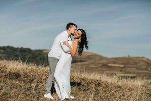 A stylish model couple in the mountains in the summer. A young boy and a girl in a white silk dress are walking on the slope against the background of the forest and mountain peaks. boy kisses a girl photo