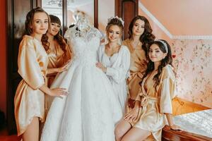 Young bridesmaids in silk robes in a hotel room. Beautiful women celebrate a bachelorette party, next to a luxurious dress of the bride in themed pajamas. photo