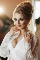 A blonde bride in a long, lacy petticoat is standing in her room, posing next to her wedding dress. Beautiful hair and make-up, open bust photo