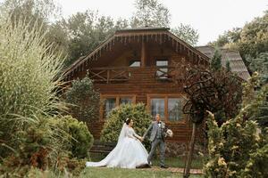 Handsome groom and charming bride stay together near modern wooden house in park. Wide-angle photo, place for text photo