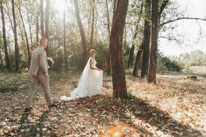 Wedding photo in nature. Bridal walk in the forest, the bride is in front of the groom, looking at him over his shoulder, smiling. Sunbeam in the photo. Couple in love.
