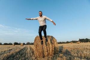 Wedding photo. Stylish groom in white shirt jumping down from hay bale and looking at camera. Bearded man. Style. Emotions photo