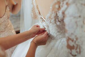 The bride's friends fasten the white voluminous dress of the bride. Rear view. Open shoulders. Beautiful hands. Long veil. Morning of the bride. photo