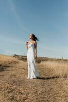 A girl with long, black, curly hair blowing in the wind is standing dressed in a white silk dress, holding a glass of champagne in her hand, against a background of blue sky and golden steppe. photo