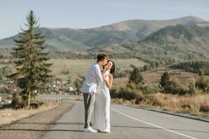 un elegante novio en un blanco camisa y un linda morena novia en un blanco vestir son caminando en un asfalto la carretera en contra el antecedentes de un bosque y montañas. Boda retrato de recién casados. foto