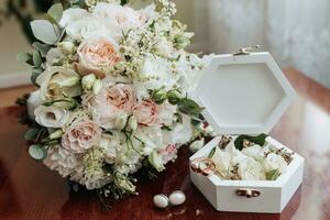 Photo of wedding accessories. Luxurious bouquet of the groom. Gold rings on a stand with flowers, white cufflinks on a wooden floor background.