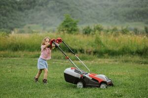un niño en botas en el formar de un juego corta césped con un cortacésped en el yarda en contra el antecedentes de montañas y niebla, el concepto de jardín herramientas foto