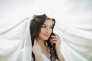 A beautiful bride with long curly hair and a delicate tiara on her head, wrapped in a white veil, gently holds her hair. Portrait of a girl. Natural makeup photo