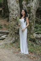 A girl with long black curly hair, standing in a white silk dress in a spring forest against a large tree, holding a fern branch in her hands photo