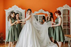 disfrutando el preparativos para el boda. damas de honor Ayudar el novia a poner en el velo y vestir y sonriente mientras en pie en el habitación foto