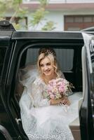 A blonde bride sits in a black car on her wedding day with a bouquet. Portrait of the bride. Beautiful makeup and hair. Magnificent white lace dress photo